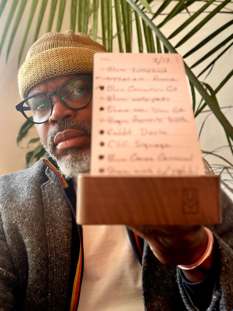 A man wearing glasses and a knit hat holds a notecard with a handwritten to-do list. The card is partially blurred, and a green plant is visible in the background.