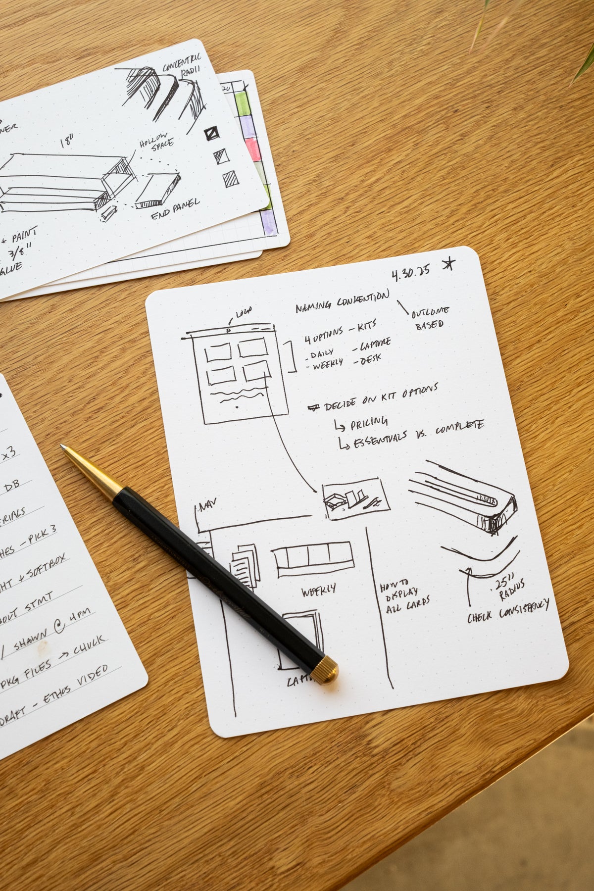 A notecard on a wooden desk displays hand-drawn sketches, notes, and a to-do list, with a black and gold pen resting on top. Another notecard with additional notes and sketches is partially visible in the background.