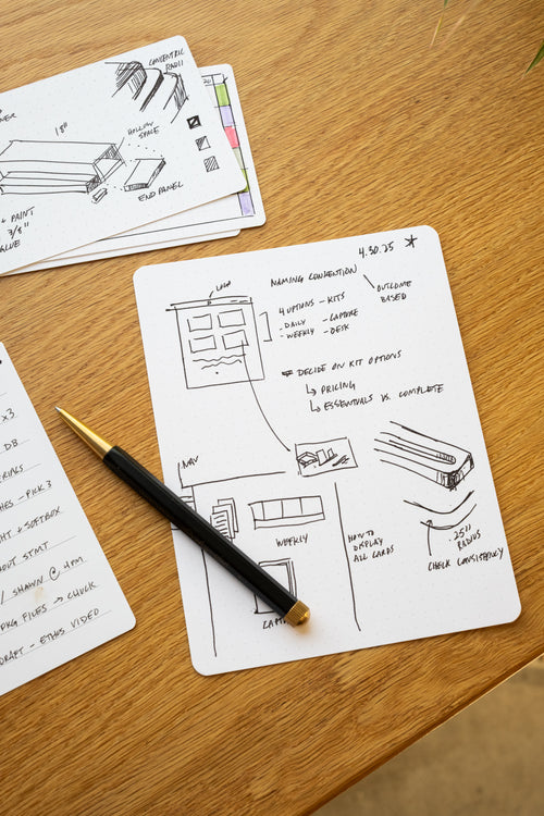 A notecard on a wooden desk displays hand-drawn sketches, notes, and a to-do list, with a black and gold pen resting on top. Another notecard with additional notes and sketches is partially visible in the background.