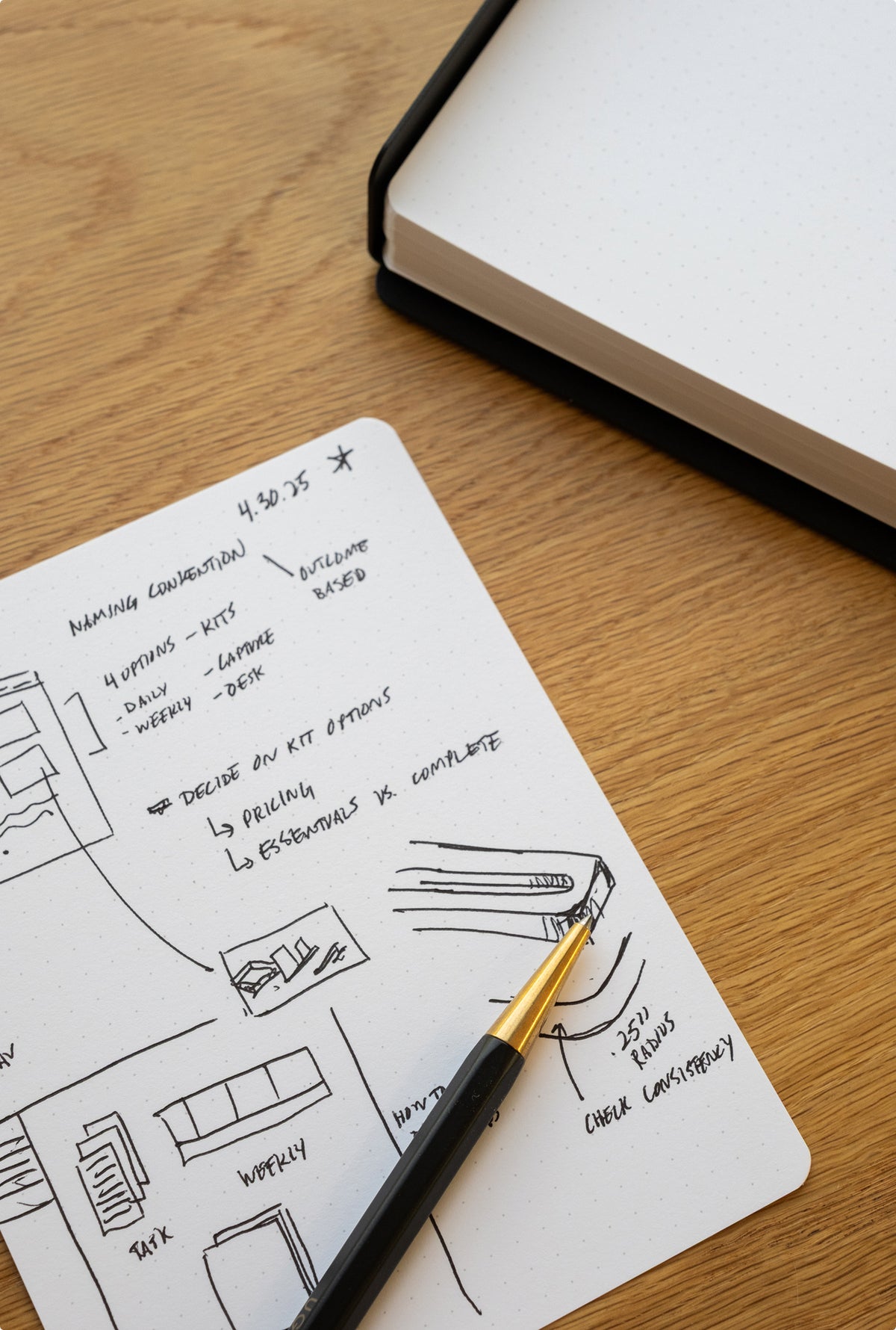 A black and gold pen rests on Analog Dot Grid Cards - Large with handwritten notes and sketches, while an open, blank dotted notebook lies nearby on a wooden desk.