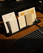 A desk with a keyboard and a black stand holding sketches, notes, and a drawing of chairs. The Analog Card Bar (Large 16" – Black), crafted from solid aluminum, sits neatly beside a desktop pen tray, organizing essentials as sunlight casts shadows across the wooden surface.