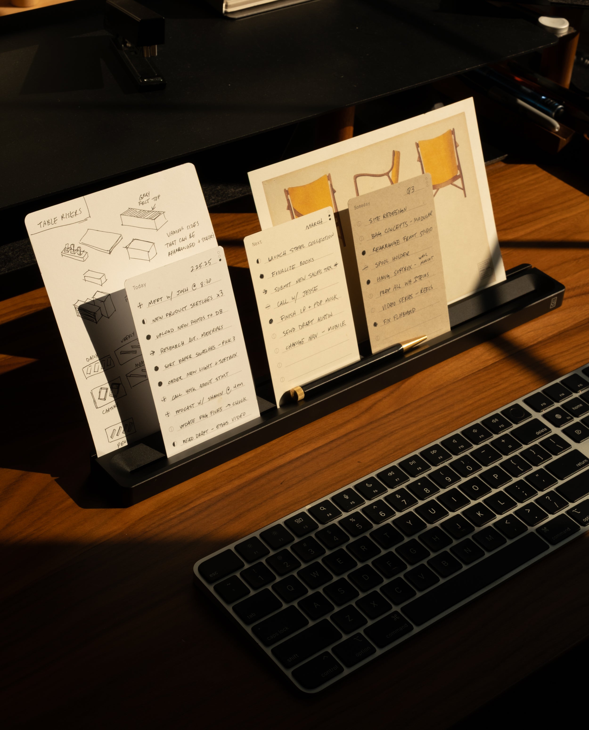 A desk with a keyboard and a black stand holding sketches, notes, and a drawing of chairs. The Analog Card Bar (Large 16" – Black), crafted from solid aluminum, sits neatly beside a desktop pen tray, organizing essentials as sunlight casts shadows across the wooden surface.