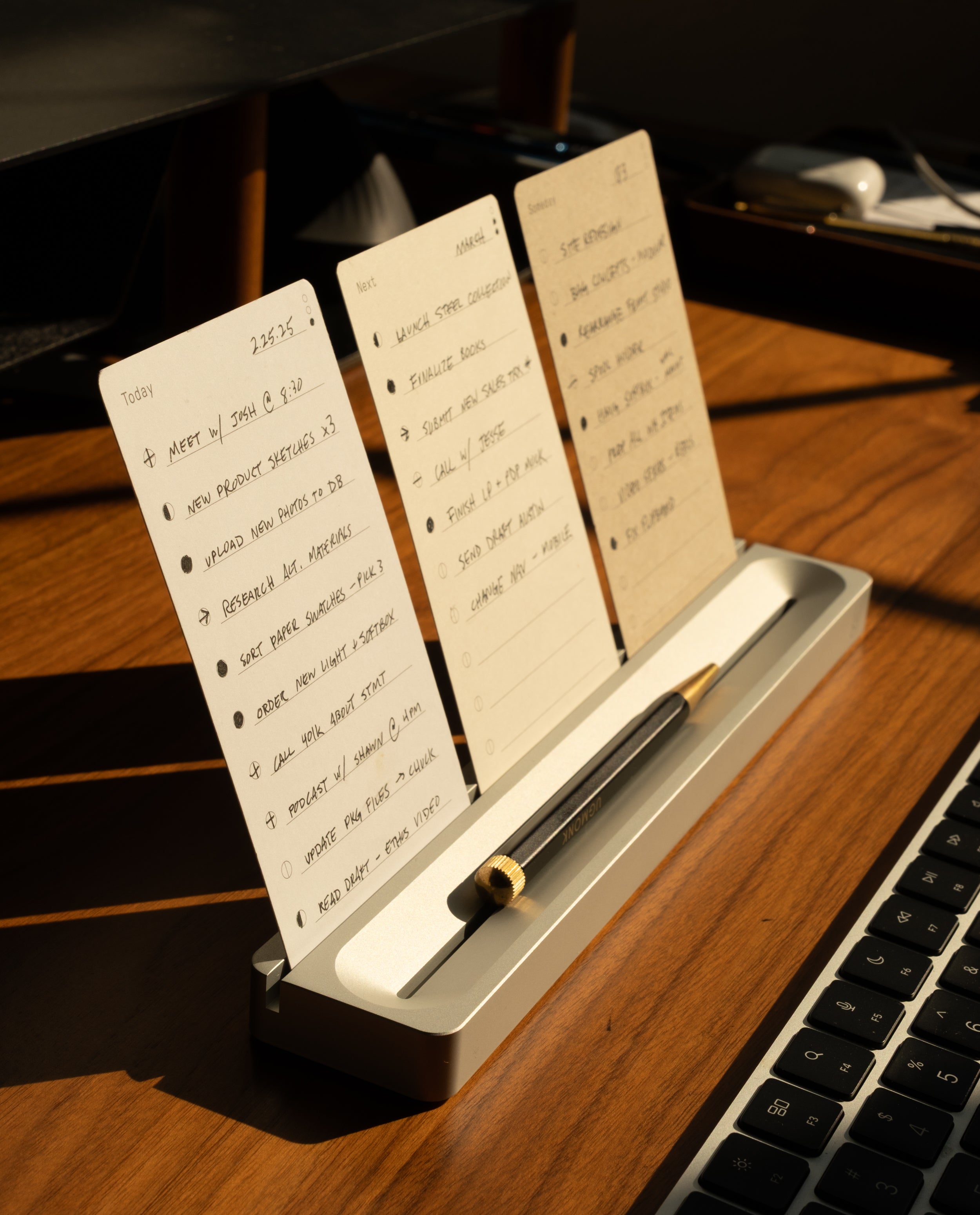 A wooden desk with sunlight illuminating the modern Analog Card Bar - Essentials Kit (Silver Aluminum), displaying handwritten lists on dot grid cards, a pen resting in the holder, and a computer keyboard nearby.