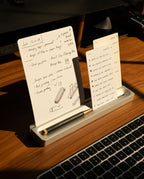 Two handwritten note cards from the Analog Card Bar - Essentials Kit (Silver Aluminum) sit in their sleek metal holder atop a wooden desk, accompanied by a black pen and part of a laptop keyboard, all illuminated by sunlight. These dot grid cards are perfect for jotting down quick notes or organizing your thoughts with clarity and style.