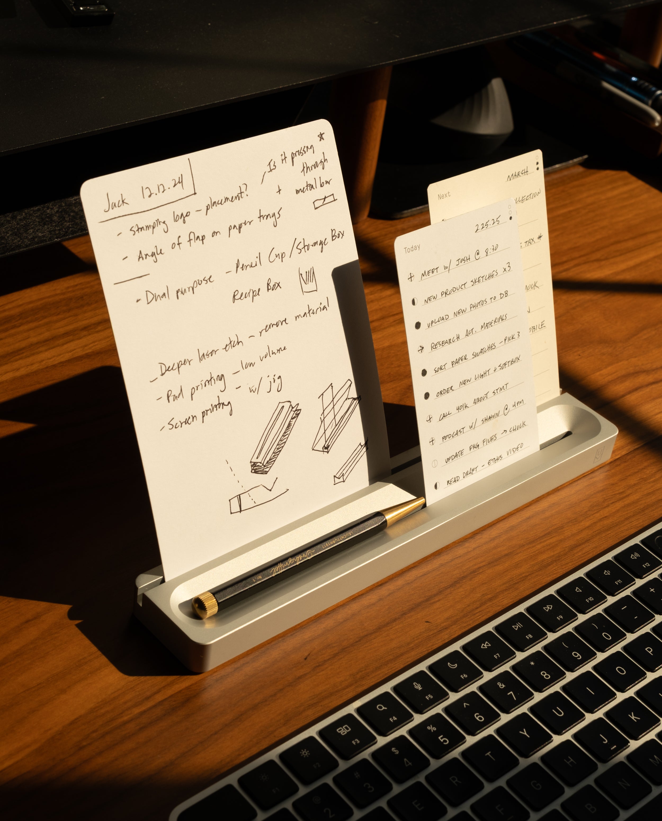 Two note cards with handwritten notes rest in the Analog Card Bar (Small 10" - Silver) solid aluminum card holder on a wooden desk, next to a black pen and in front of a laptop keyboard, illuminated by warm sunlight.