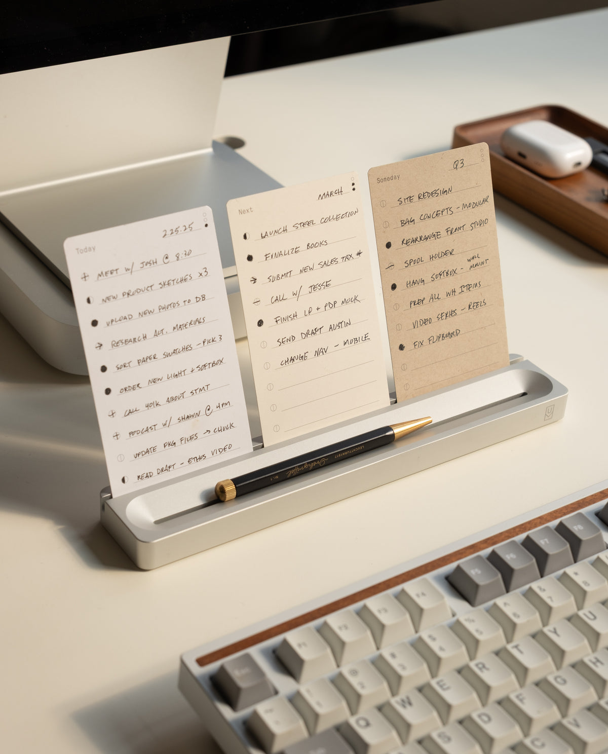 Three notecards featuring handwritten to-do lists are displayed upright on the Analog Card Bar - Essentials Kit (Silver Aluminum), with a black pen resting in front. A keyboard and a wooden tray holding wireless earbuds are also visible on the tidy desk, showcasing the efficient use of dot grid cards for organizing tasks.