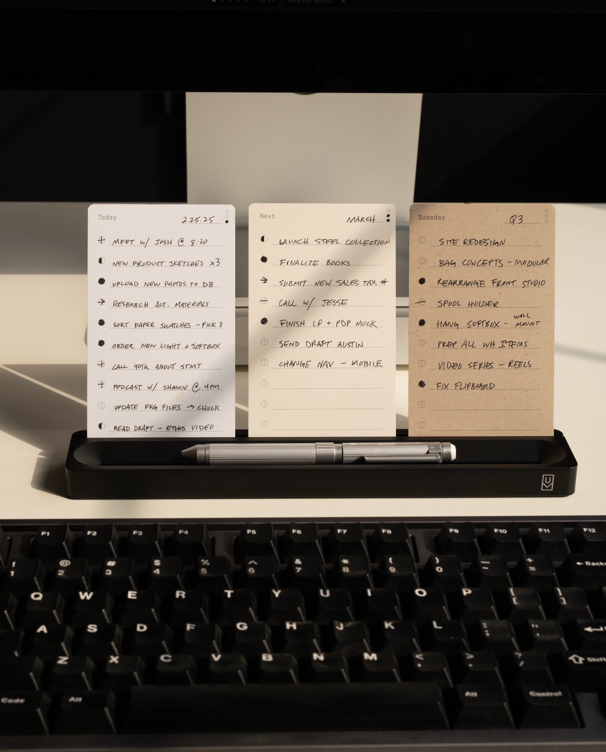 Three notecards with handwritten task lists are propped on the Analog Card Bar (Small 10" - Black), paired with a silver pen, all set behind a black keyboard on a warmly lit desk.