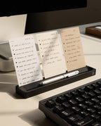 The Analog Card Bar (Small 10" - Black) holds three notecards upright on a desk beside a keyboard and monitor, with a silver pen resting in the holder for an efficient and organized workspace setup.