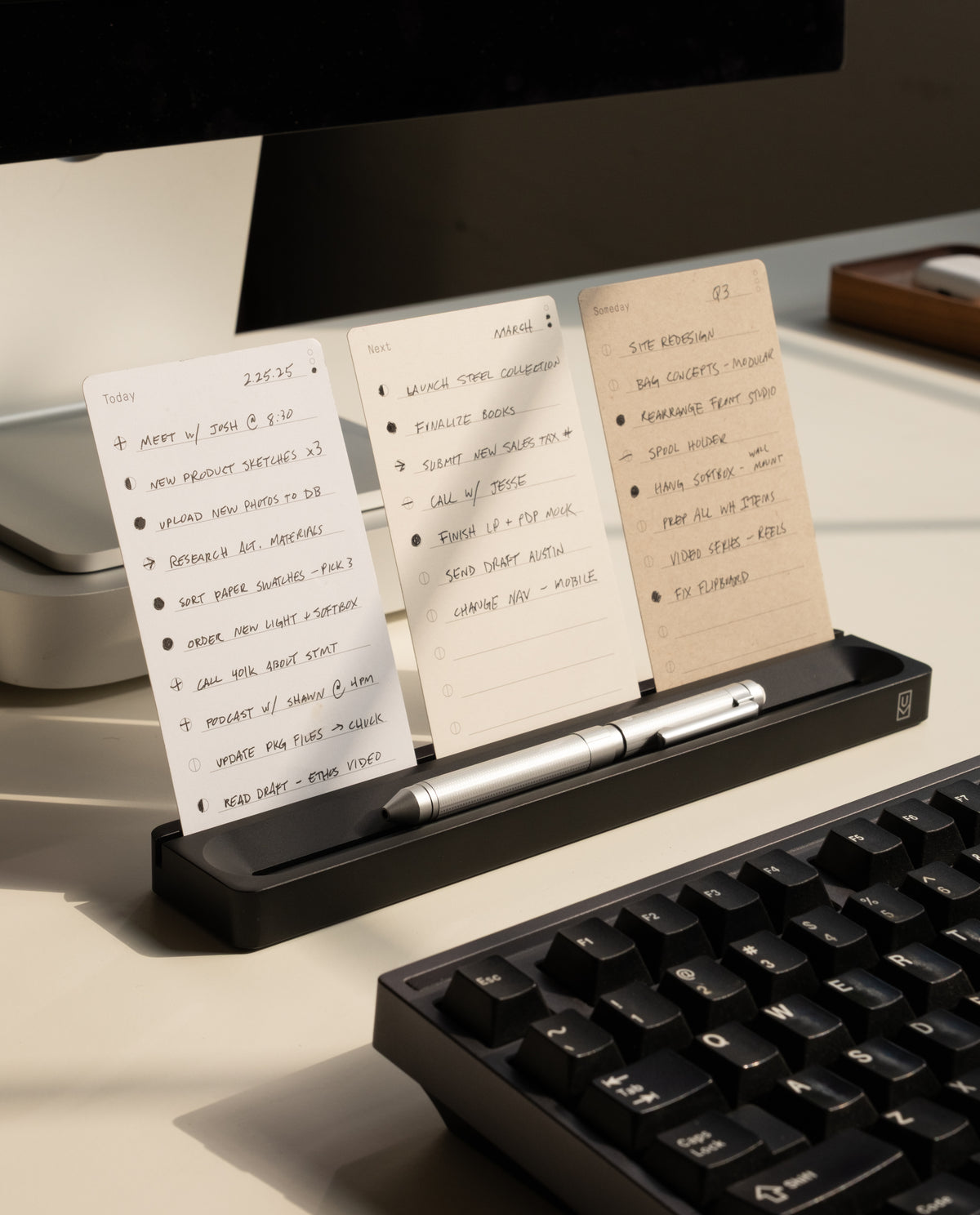 The Analog Card Bar (Small 10" - Black) holds three notecards upright on a desk beside a keyboard and monitor, with a silver pen resting in the holder for an efficient and organized workspace setup.