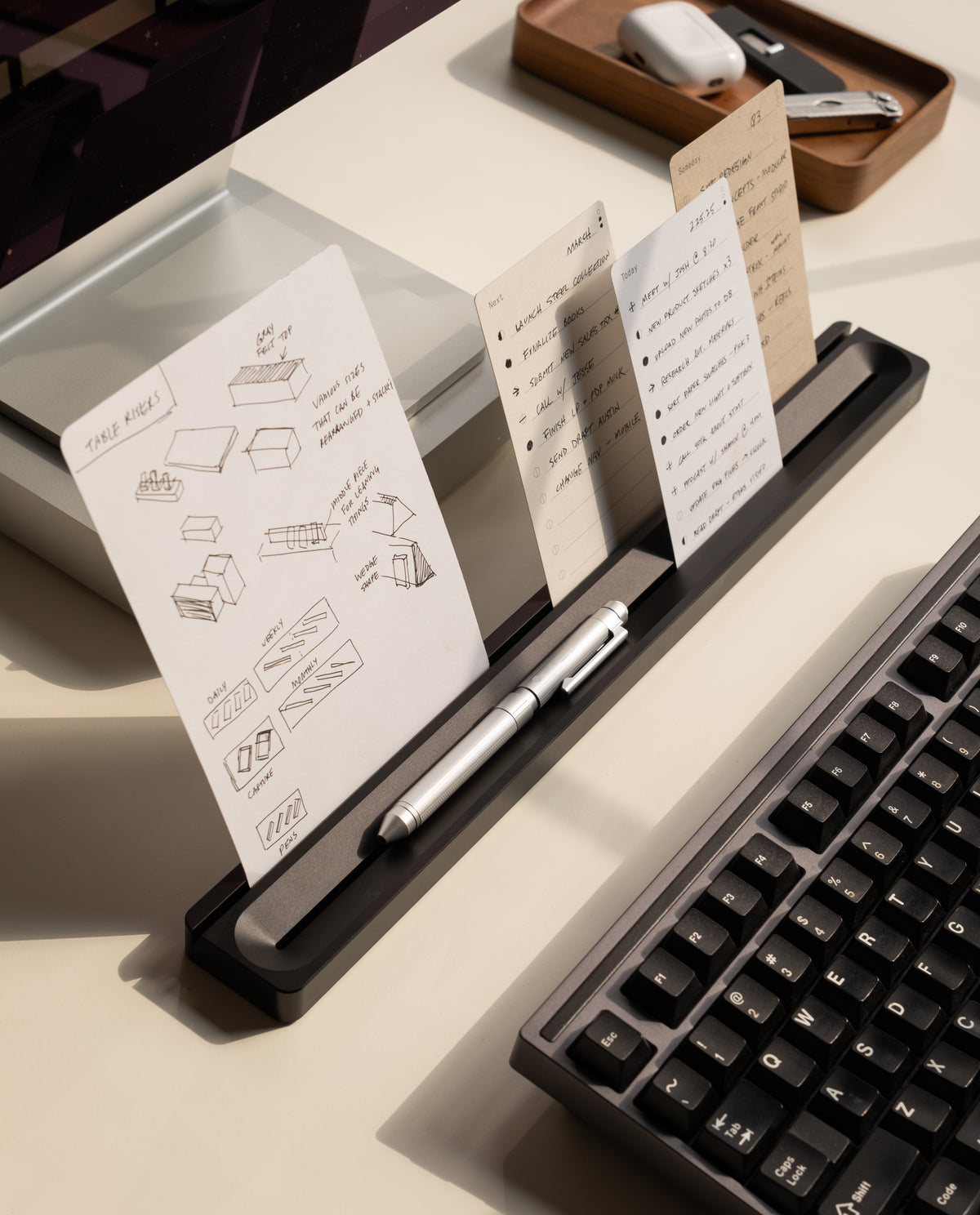 A desktop organizer holds pens and several sheets of notes, sketches, and diagrams next to a computer keyboard. The Analog Card Bar (Large 16" – Black), made of solid aluminum, keeps business cards neatly displayed alongside the desktop pen tray, while a tray with AirPods and a stapler sits in the background.