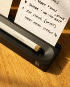 A white UGMONK pen rests in a black Analog Card Bar (Large 16") on a wooden surface, with handwritten notes on white cards and a solid aluminum card holder visible in the background.