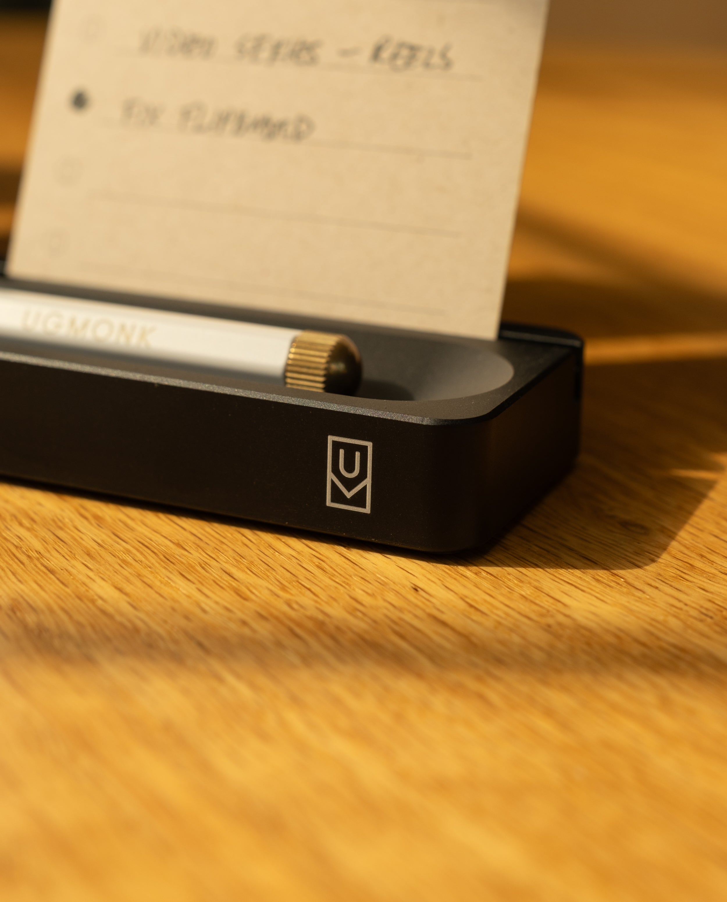 A close-up of the Analog Card Bar (Small 10" - Black), a sleek solid aluminum organizer holding a pen and an index card with handwritten notes. Sunlight creates soft shadows on the black desk accessory atop a wooden surface.