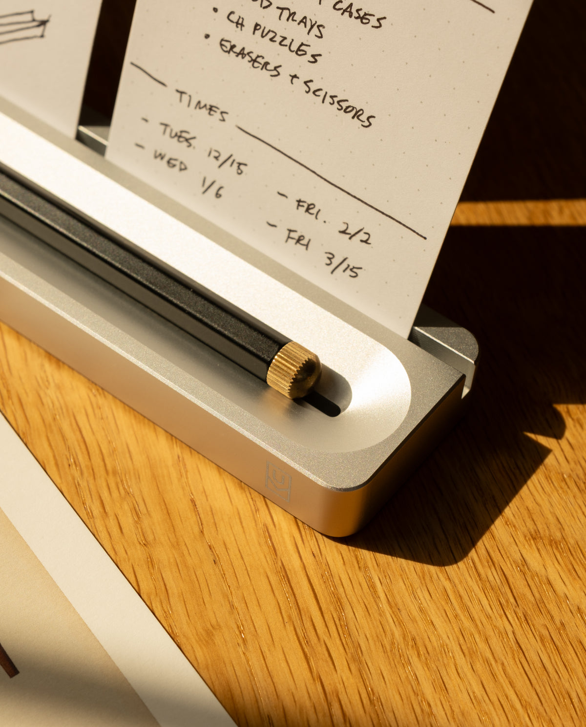 A close-up of the Analog Card Bar - Essentials Kit in Silver Aluminum displays a black pencil with a gold eraser cap resting in the sleek silver holder, surrounded by dot grid cards and notepads featuring handwritten notes and dates, all arranged on a wooden surface illuminated by warm sunlight.