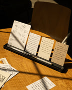 Four small notepads with sketches and handwritten notes stand upright in an Analog Card Bar (Large 16" – Black) solid aluminum card holder on a wooden table, with pens neatly arranged in a desktop pen tray in front. Sunlight casts shadows across the scene, while a chair and additional paper are visible in the background.