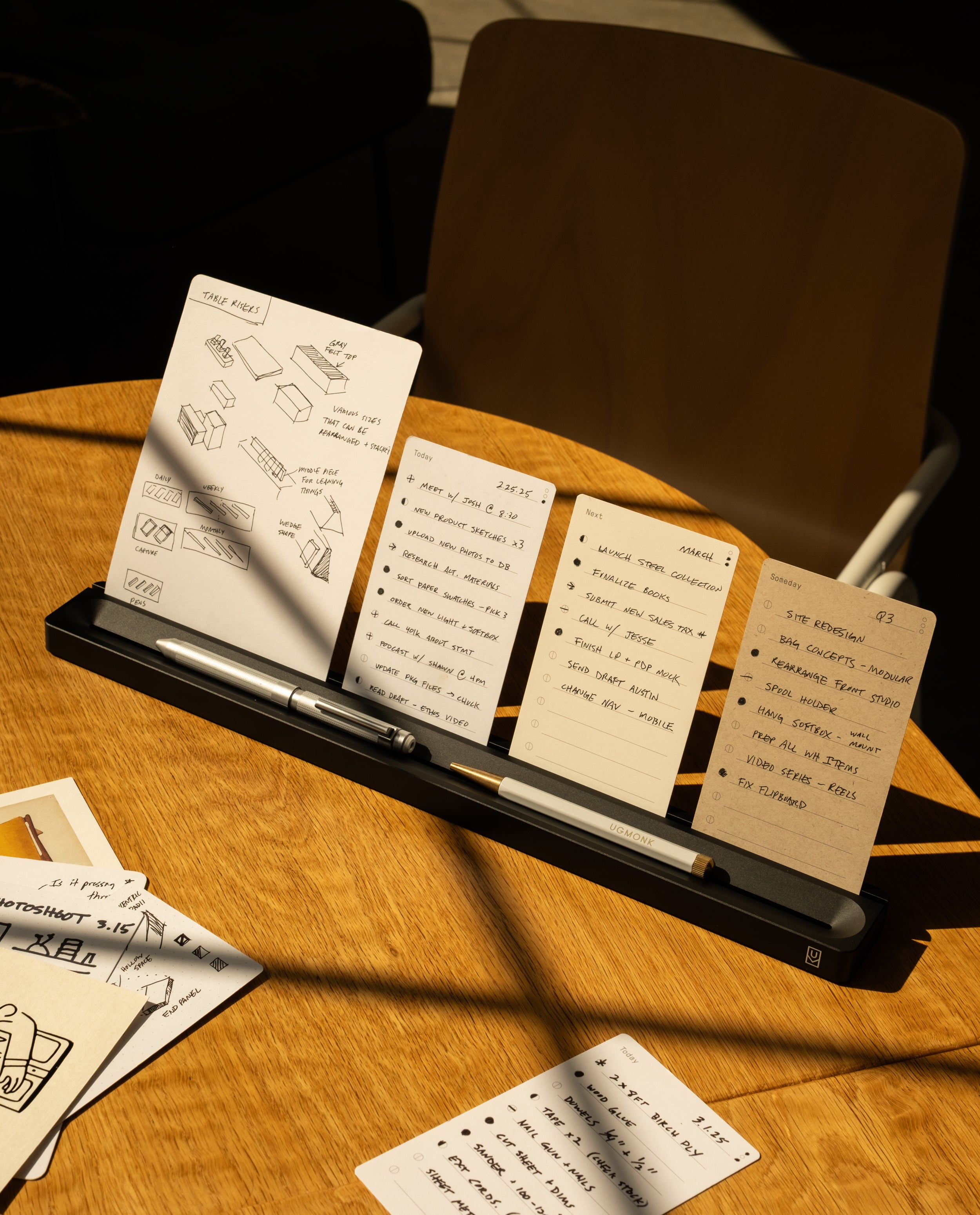Four small notepads with sketches and handwritten notes stand upright in an Analog Card Bar (Large 16" – Black) solid aluminum card holder on a wooden table, with pens neatly arranged in a desktop pen tray in front. Sunlight casts shadows across the scene, while a chair and additional paper are visible in the background.