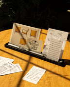 On a sunlit wooden table, the Analog Card Bar (Large 16" – Black) organizes a pen, a card that says THANKS, and a photo of chairs, with handwritten notes neatly arranged nearby. The solid aluminum design serves as both a stylish desktop pen tray and card holder, while additional notes and drawings are spread across the surface for easy access.