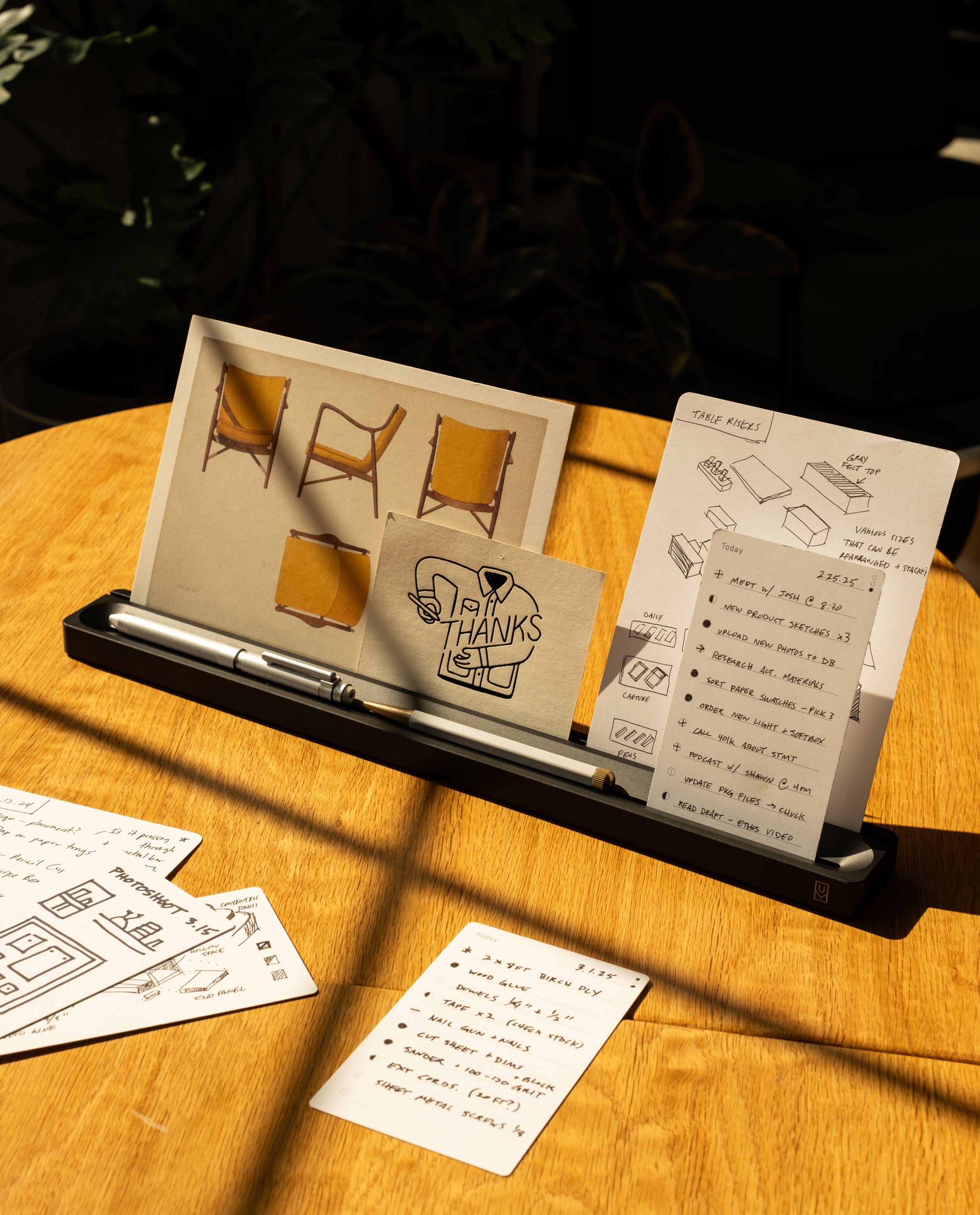 On a sunlit wooden table, the Analog Card Bar (Large 16" – Black) organizes a pen, a card that says THANKS, and a photo of chairs, with handwritten notes neatly arranged nearby. The solid aluminum design serves as both a stylish desktop pen tray and card holder, while additional notes and drawings are spread across the surface for easy access.