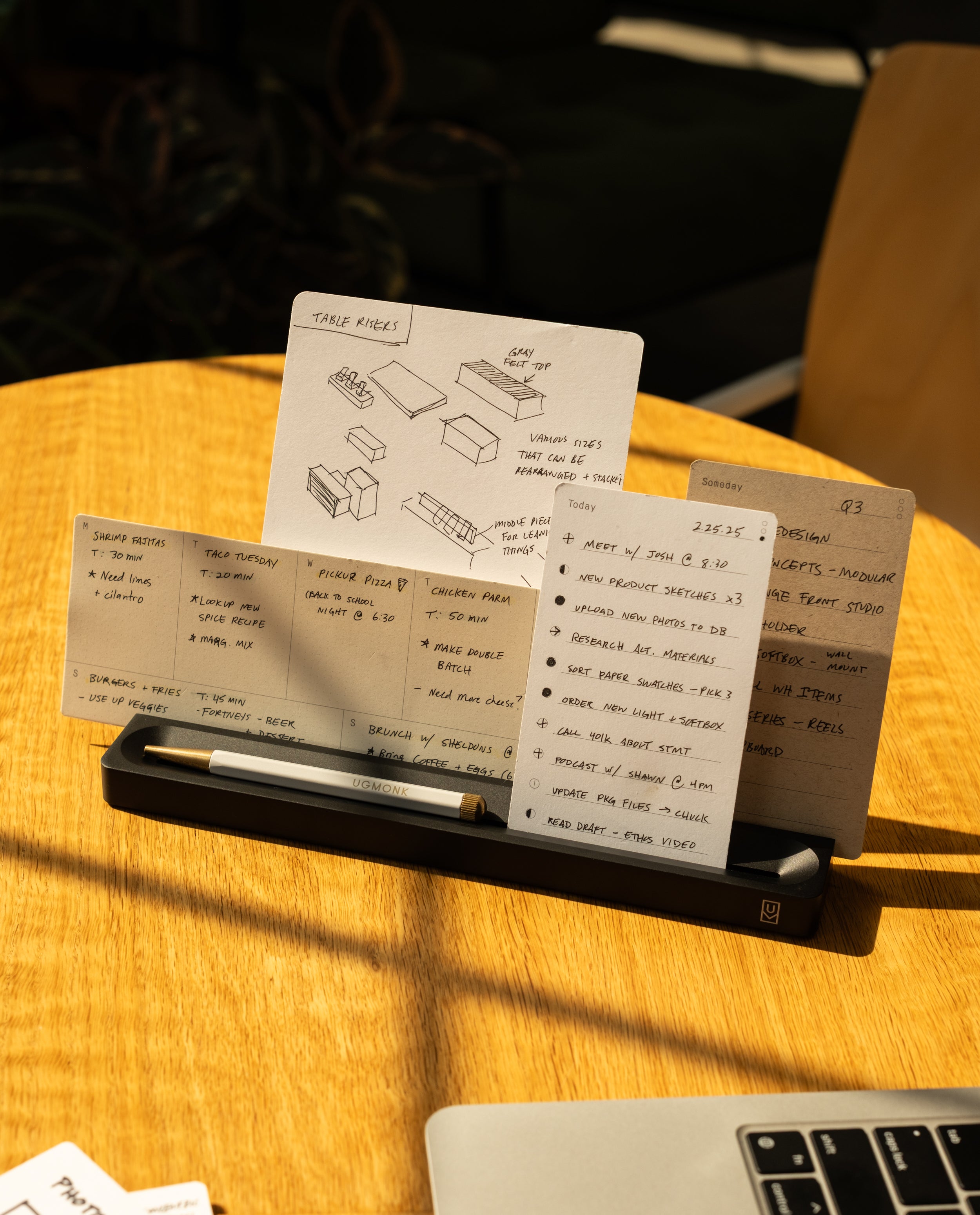 The Analog Card Bar (Small 10" - Black) neatly organizes three notecards with handwritten notes and diagrams, while a pen rests in its tray. Part of a laptop sits nearby on a sunlit wooden table.
