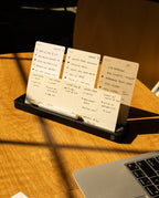 The Analog Card Bar (Small 10" - Black) holds neatly arranged handwritten note cards with to-do lists and notes beside a laptop on a wooden table, all illuminated by warm sunlight.