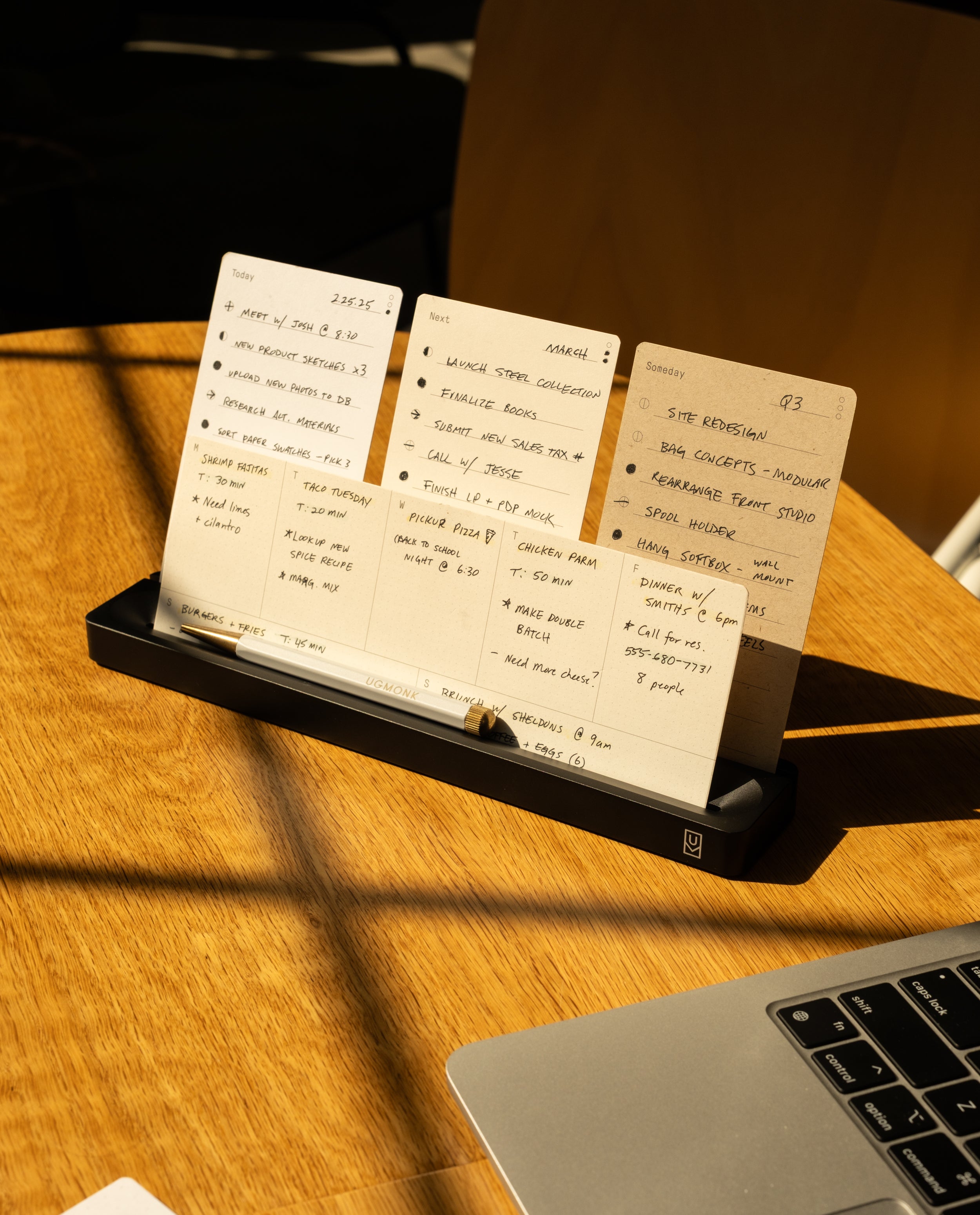 The Analog Card Bar (Small 10" - Black) holds neatly arranged handwritten note cards with to-do lists and notes beside a laptop on a wooden table, all illuminated by warm sunlight.