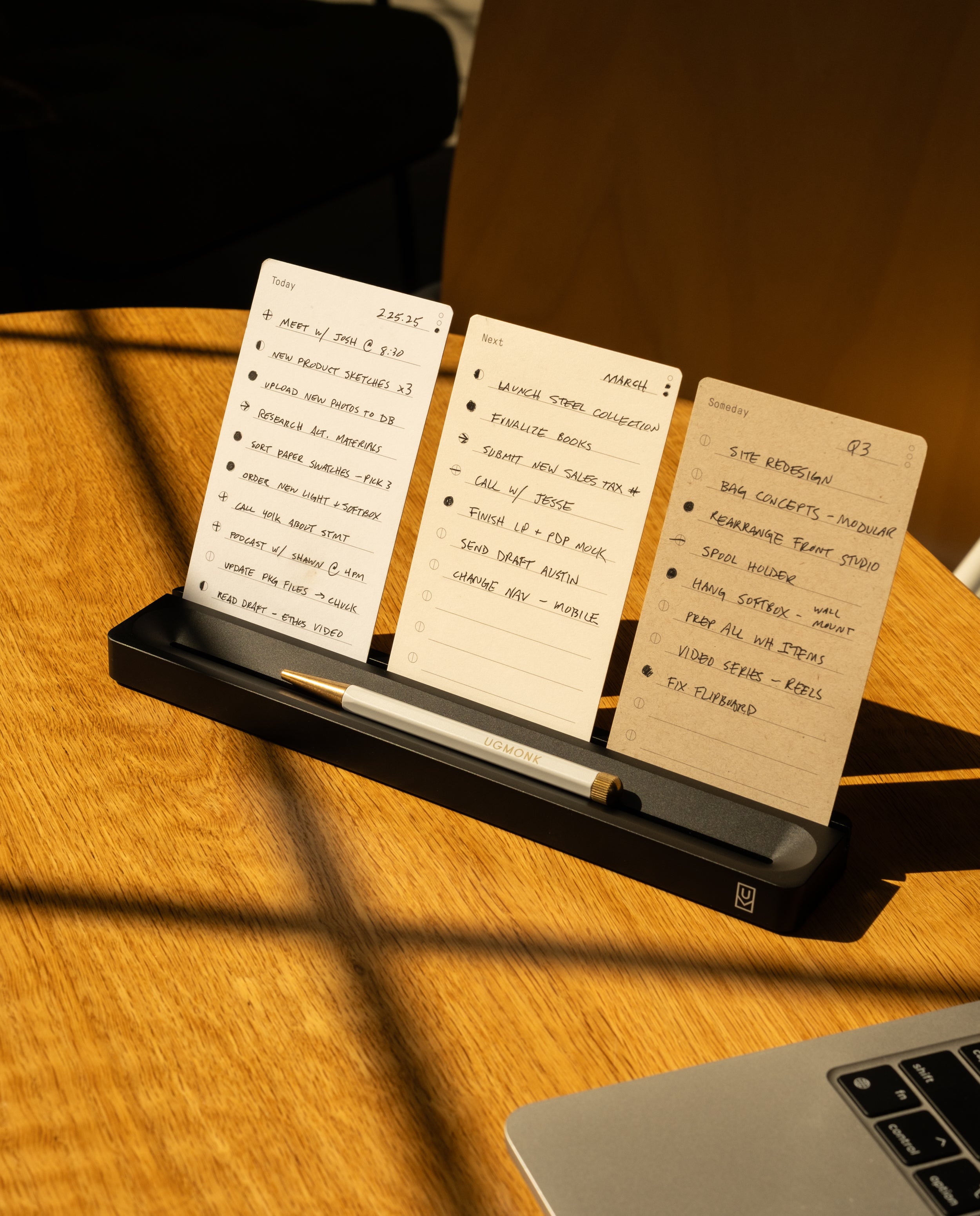 Three notecards with handwritten to-do lists for different days are displayed upright in a black holder on a wooden desk, with a pen in front. Warm sunlight casts shadows across the scene.