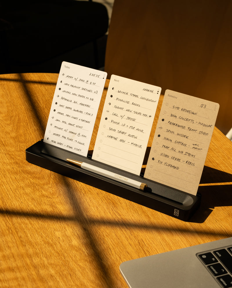 Three notecards with handwritten to-do lists for different days are displayed upright in a black holder on a wooden desk, with a pen in front. Warm sunlight casts shadows across the scene.