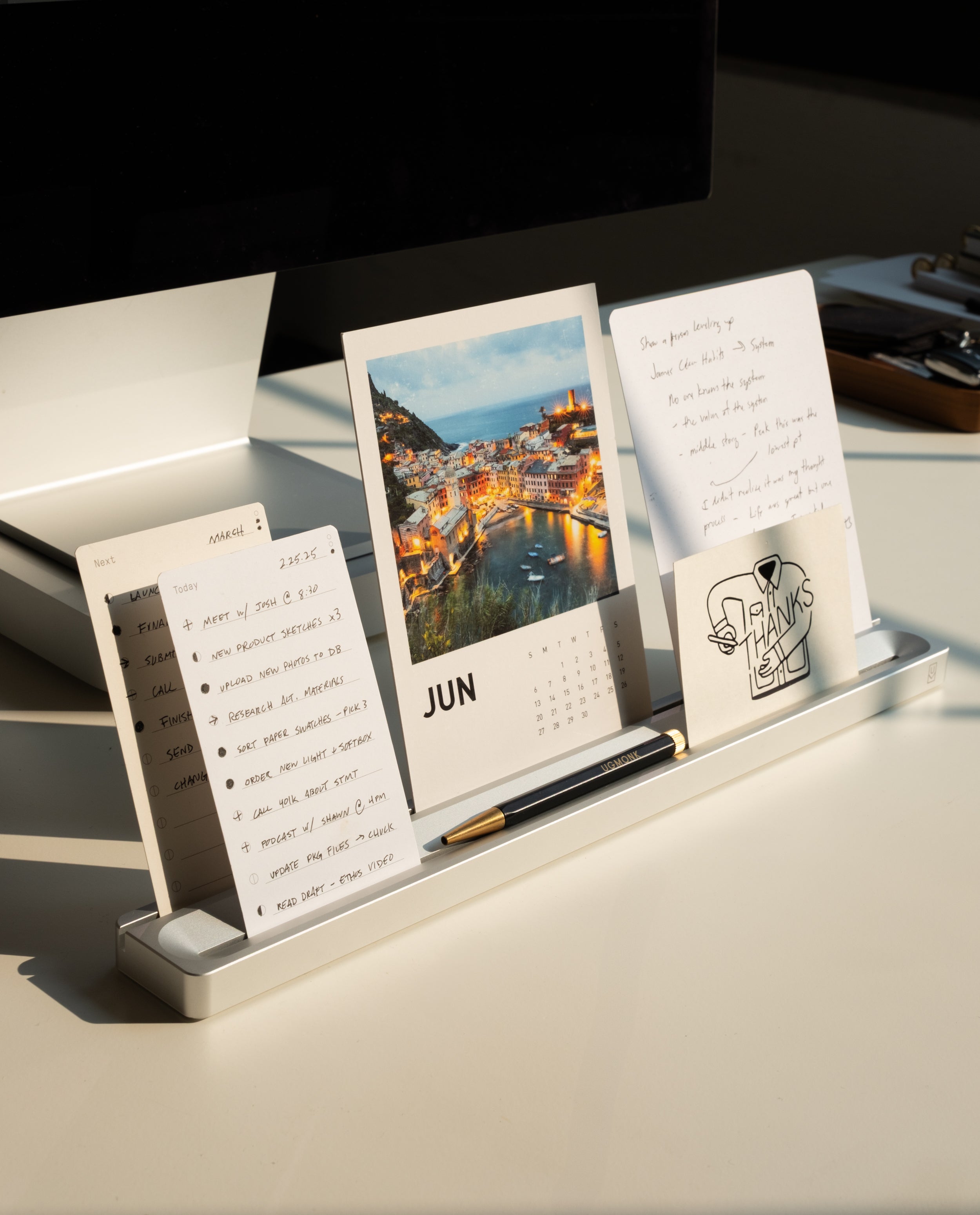 A solid aluminum Analog Card Bar (Large 16" – Silver) holds a calendar turned to June, handwritten notes, a “THANKS!” card perched in its display slot, and a pen nestled in the built-in tray—all illuminated by sunlight on a white desk near a computer monitor.
