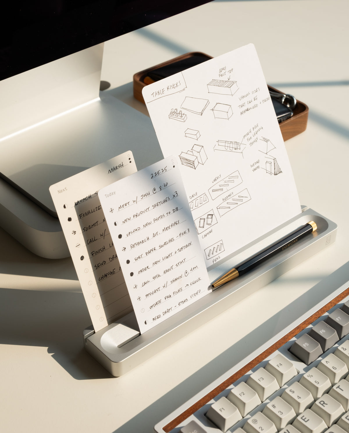 Two sheets from the Analog Card Bar - Essentials Kit (Silver Aluminum) rest on a stand by a keyboard—one features task cards, while the other displays hand-drawn box diagrams. A pen lies in front, with sunlight casting shadows across the workspace.