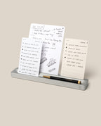 A silver Aluminum Analog Card Bar - Essentials Kit holds a black pen and three dot grid cards featuring handwritten notes, sketches, and to-do lists. The task cards stand upright in the slots of the Card Bar against a plain beige background.
