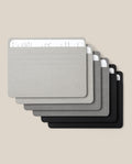 Six Analog Card Sleeves - Large (Monochromatic) in gradient shades from gray to black, each serving as a stylish organizer for stacks of handwritten white cards, are arranged in a slightly fanned, overlapping layout on a beige background.