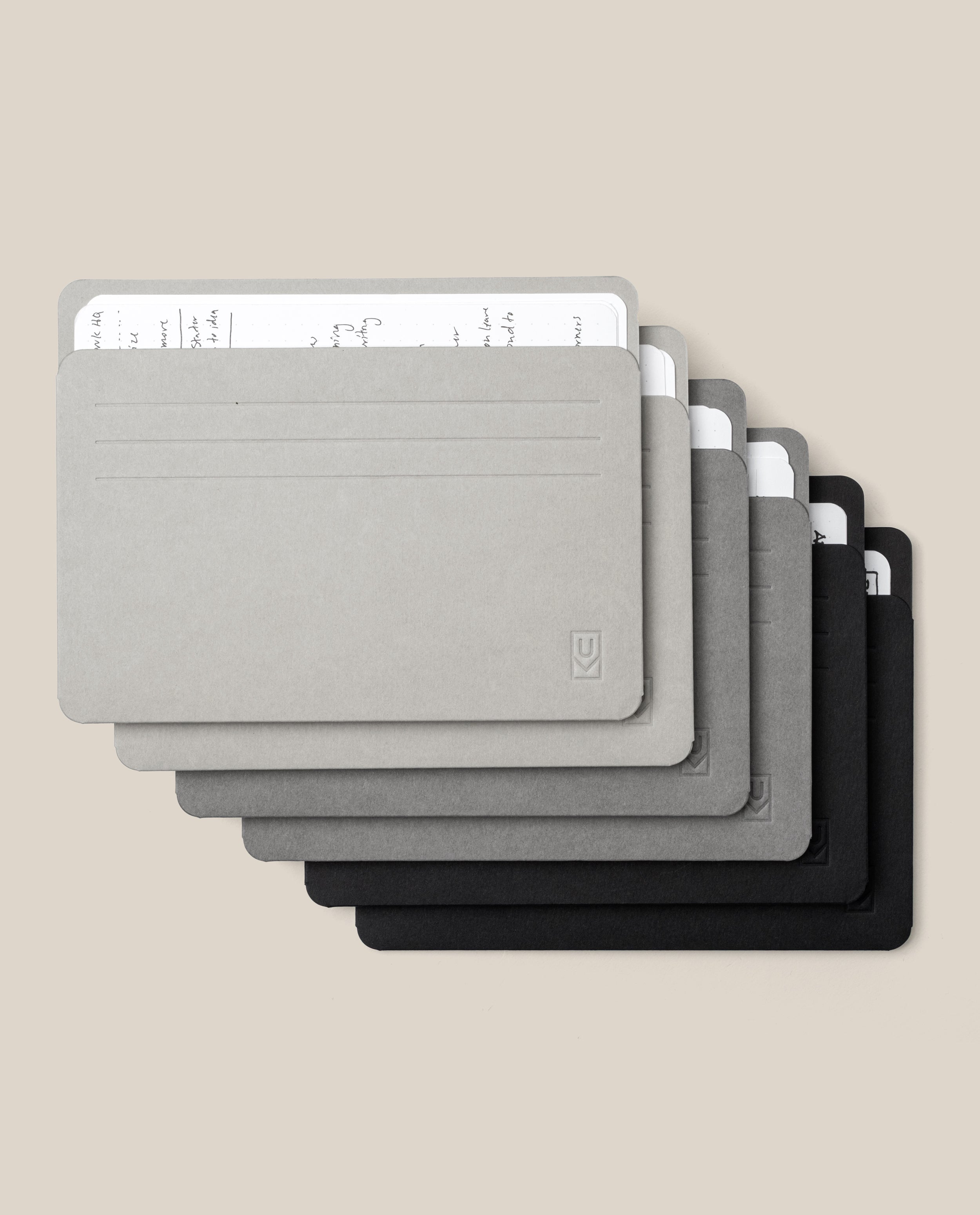 Six Analog Card Sleeves - Large (Monochromatic) in gradient shades from gray to black, each serving as a stylish organizer for stacks of handwritten white cards, are arranged in a slightly fanned, overlapping layout on a beige background.