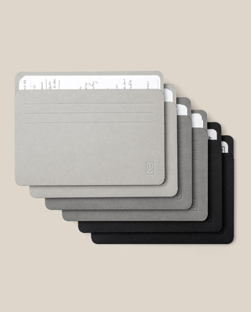 Six Analog Card Sleeves - Large (Monochromatic) in gradient shades from gray to black, each serving as a stylish organizer for stacks of handwritten white cards, are arranged in a slightly fanned, overlapping layout on a beige background.