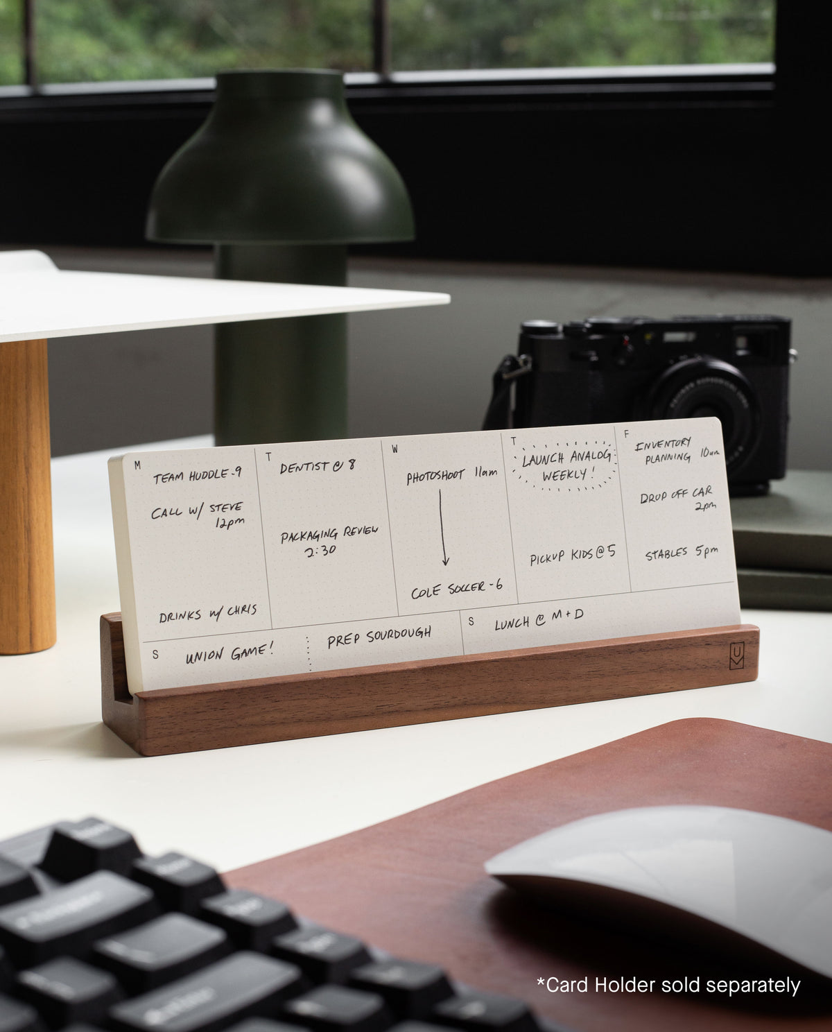 An Analog Weekly Card from our kit is displayed in a wooden holder on a desk, organized with tasks and appointments. A camera and greenery are blurred in the background. Text reads: "*Card Holder sold separately.