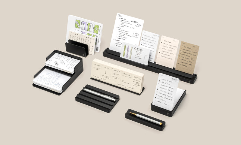 Several black desk organizers hold small cards, notepads, and pens arranged neatly on a light beige surface. The cards contain handwritten notes, sketches, and checklists in different languages.