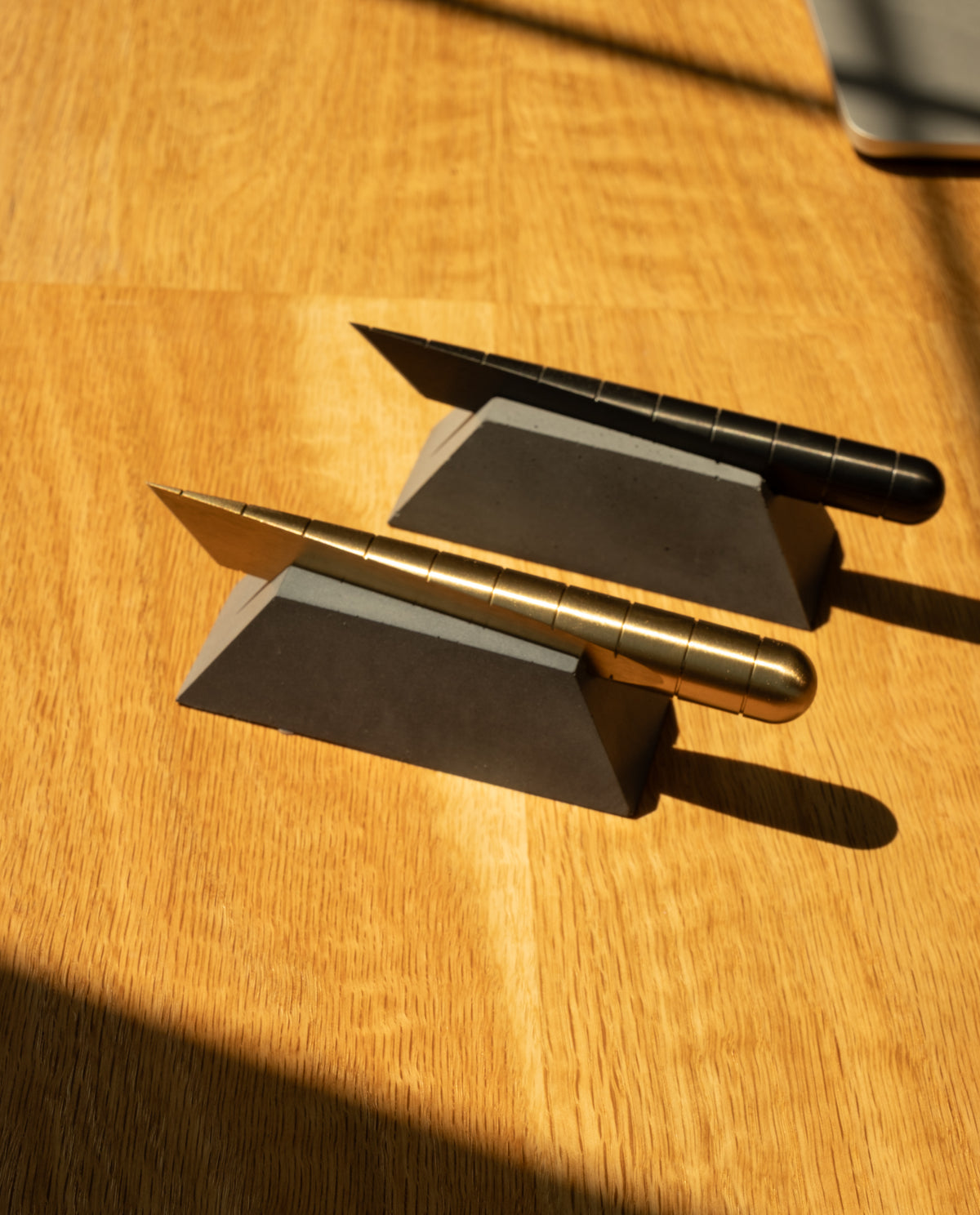 A gold and a black metallic pen rest diagonally on Craighill Desk Knife Plinths (Slate), triangular cast concrete holders arranged on a wooden surface, with sunlight casting shadows across the scene.