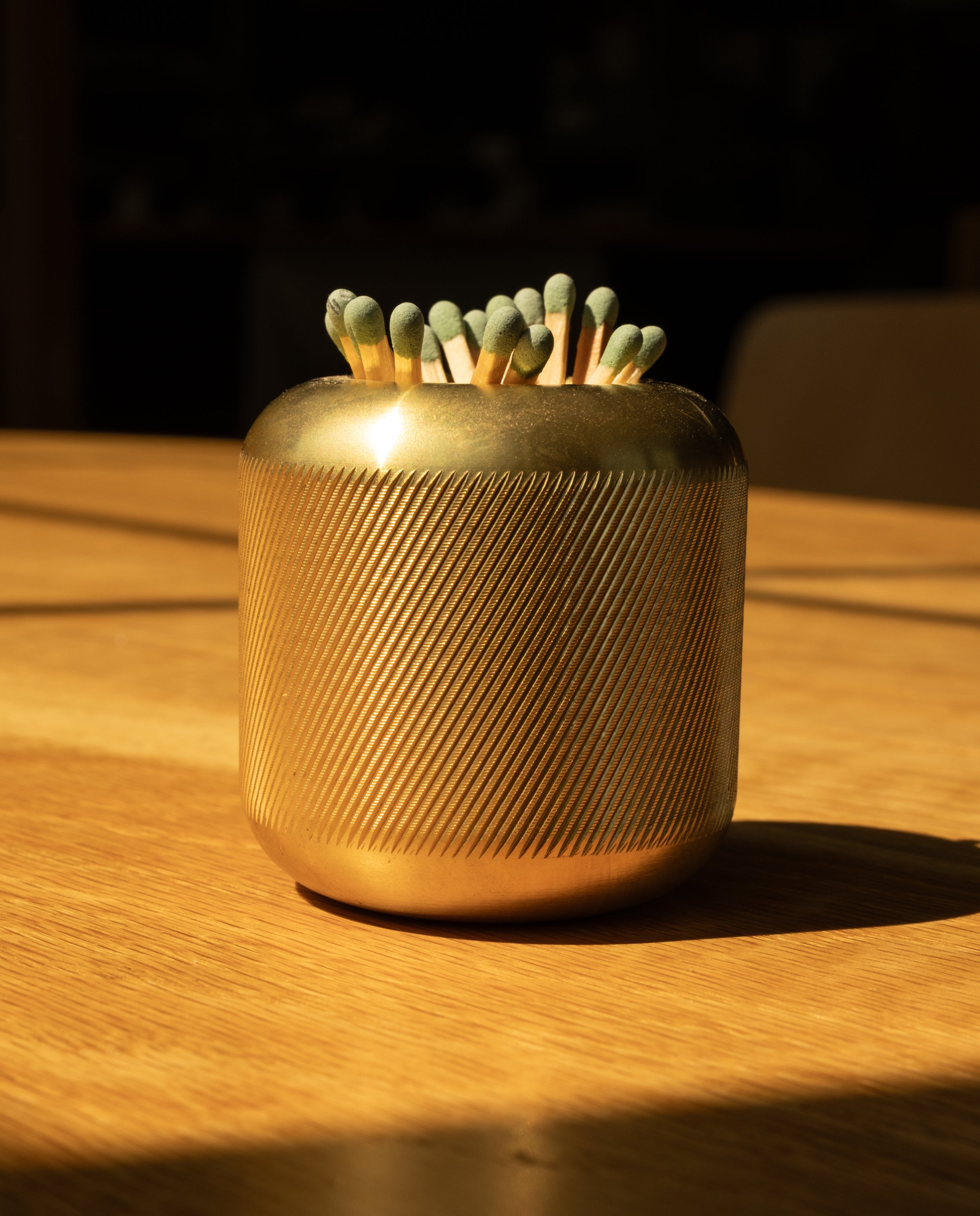The Craighill Match Striker (Brass), crafted from golden, textured solid brass, holds dozens of green-tipped strike anywhere matches and rests elegantly on a sunlit wooden surface.