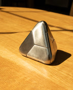 The Craighill Tetra Puzzle (Stainless Steel), a striking metallic triangular brainteaser, rests on a sunlit wooden table, casting sharp shadows. Its gleaming stainless steel surface catches the light, accentuating the distinct geometric segments on each side—an eye-catching mechanical puzzle from Craighill.