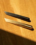 Two Craighill Desk Knives (Vapor Black) are placed parallel on a wooden surface, sunlight casting a shadow across the lower left corner. Each sleek blade—reminiscent of a Japanese kiridashi knife—serves as both a reliable desk accessory and a precise stainless steel cutting tool.