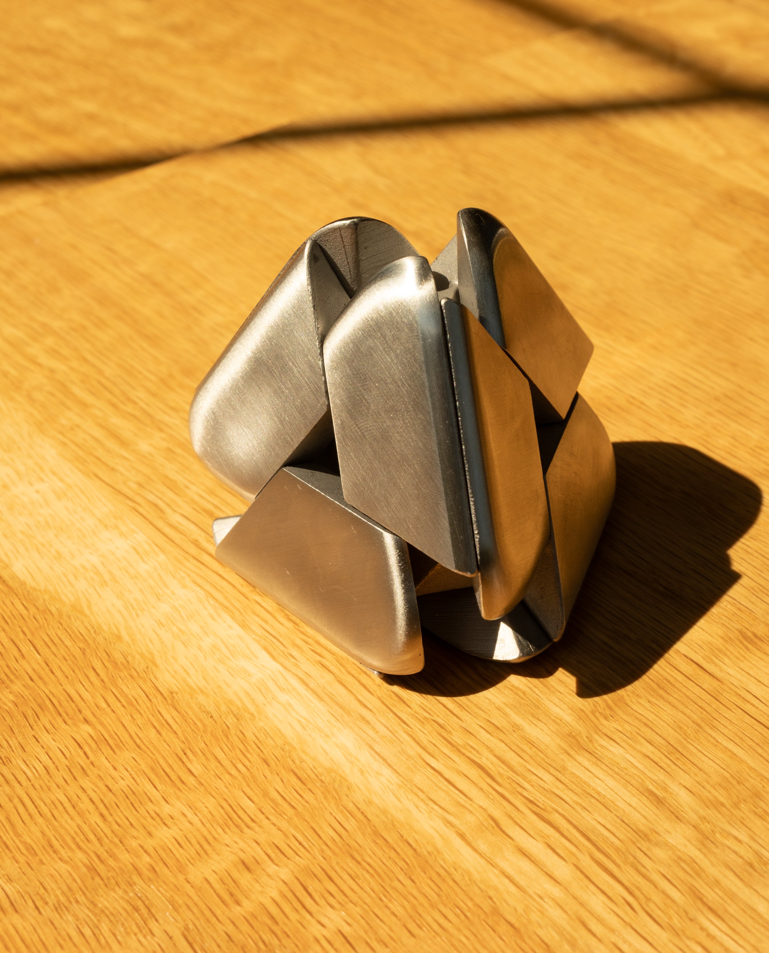 A metallic abstract sculpture with interlocking geometric shapes sits on a sunlit wooden surface, casting soft shadows—a striking piece reminiscent of the Craighill Tetra Puzzle in stainless steel.