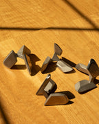 Six abstract metal sculptures rest on a wooden surface, sunlight casting angled shadows across them—each form reminiscent of the Craighill Tetra Puzzle in stainless steel, elegantly blending artistry with mechanical intrigue.