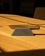 A Craighill Desk Knife with a gold handle rests on a Craighill Desk Knife Plinth (Slate), both positioned on a wooden table where sunlight casts shadows across the surface.