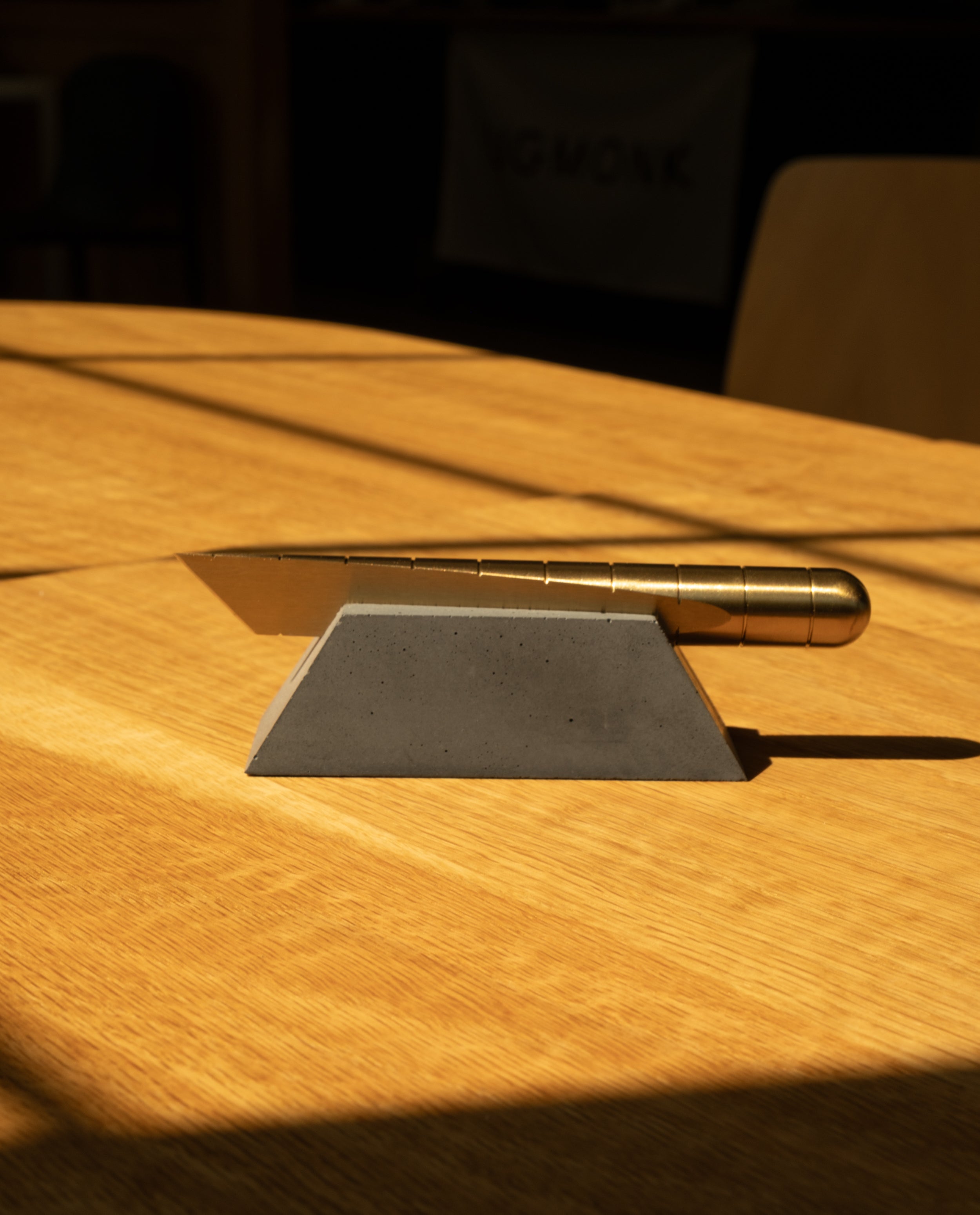 A Craighill Desk Knife with a gold handle rests on a Craighill Desk Knife Plinth (Slate), both positioned on a wooden table where sunlight casts shadows across the surface.