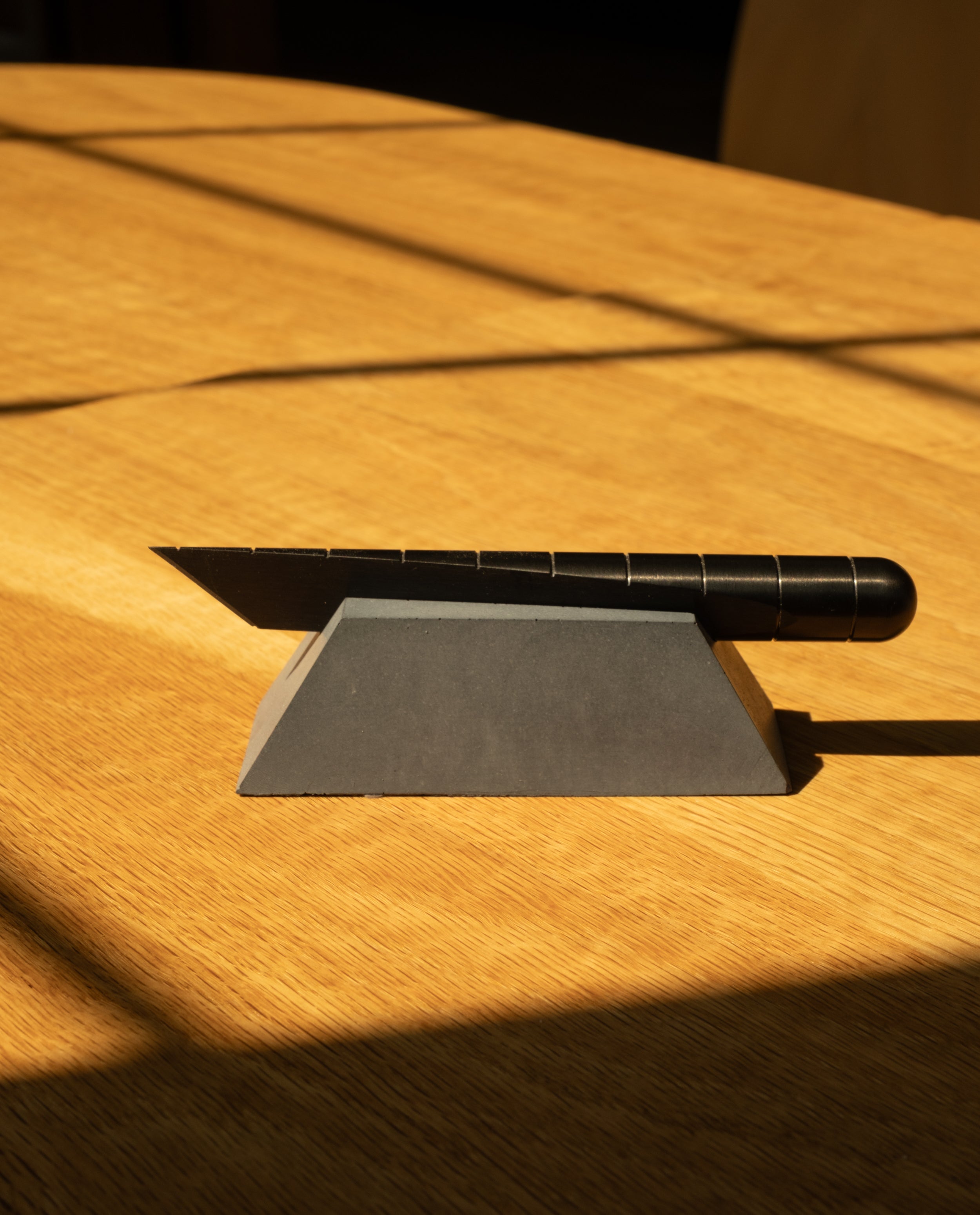 A utility knife with a black handle sits on the Craighill Desk Knife Plinth (Slate) atop a wooden surface, with sunlight casting shadows across the scene.