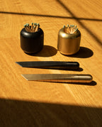 Two metal match strikers—one black, one gold—hold green-tipped matches. In front of them, two metal utility knives, including the Craighill Desk Knife in sleek brass and an elegant multipurpose cutting tool in black and gold, rest on a wooden surface with dramatic light and shadow.