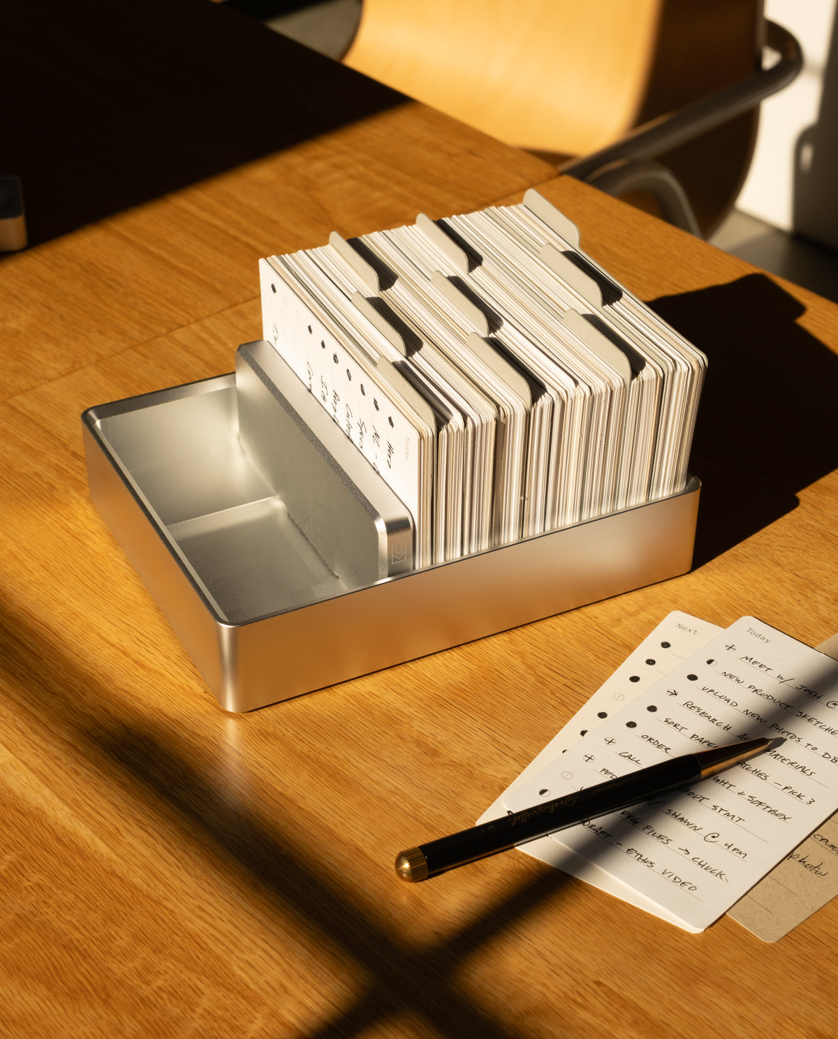 An Analog Archive (Silver Aluminum) card holder containing index cards sits on a wooden desk beside a handwritten to-do list and a pen.