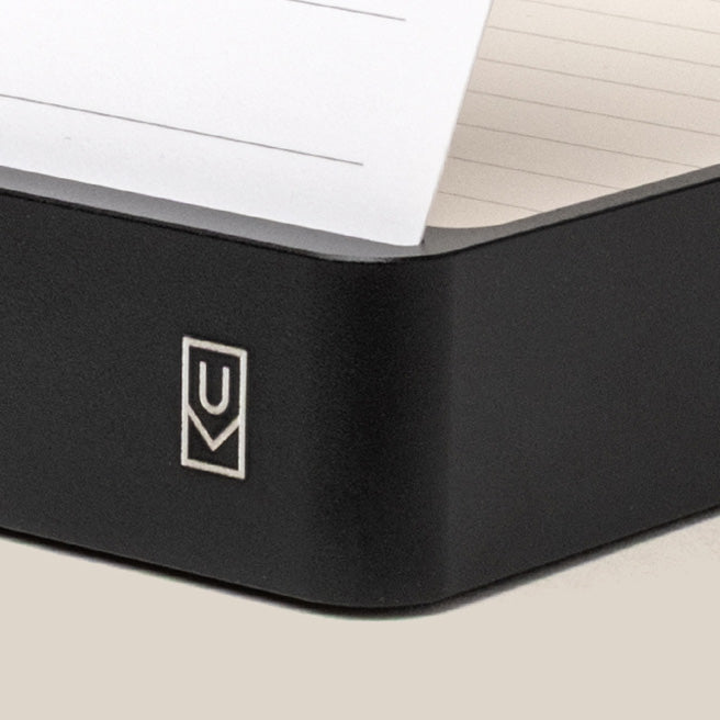 A close-up of a black notebook corner with a white logo and lined pages, one of which is slightly lifted.