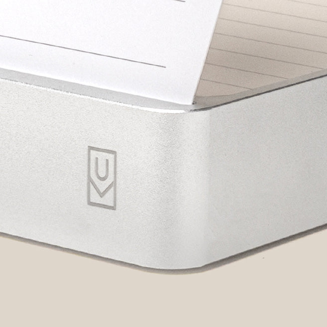 A close-up of a silver note card holder with a stack of lined index cards inside. A portion of a card is visible at the top. The holder has a subtle logo on its corner.