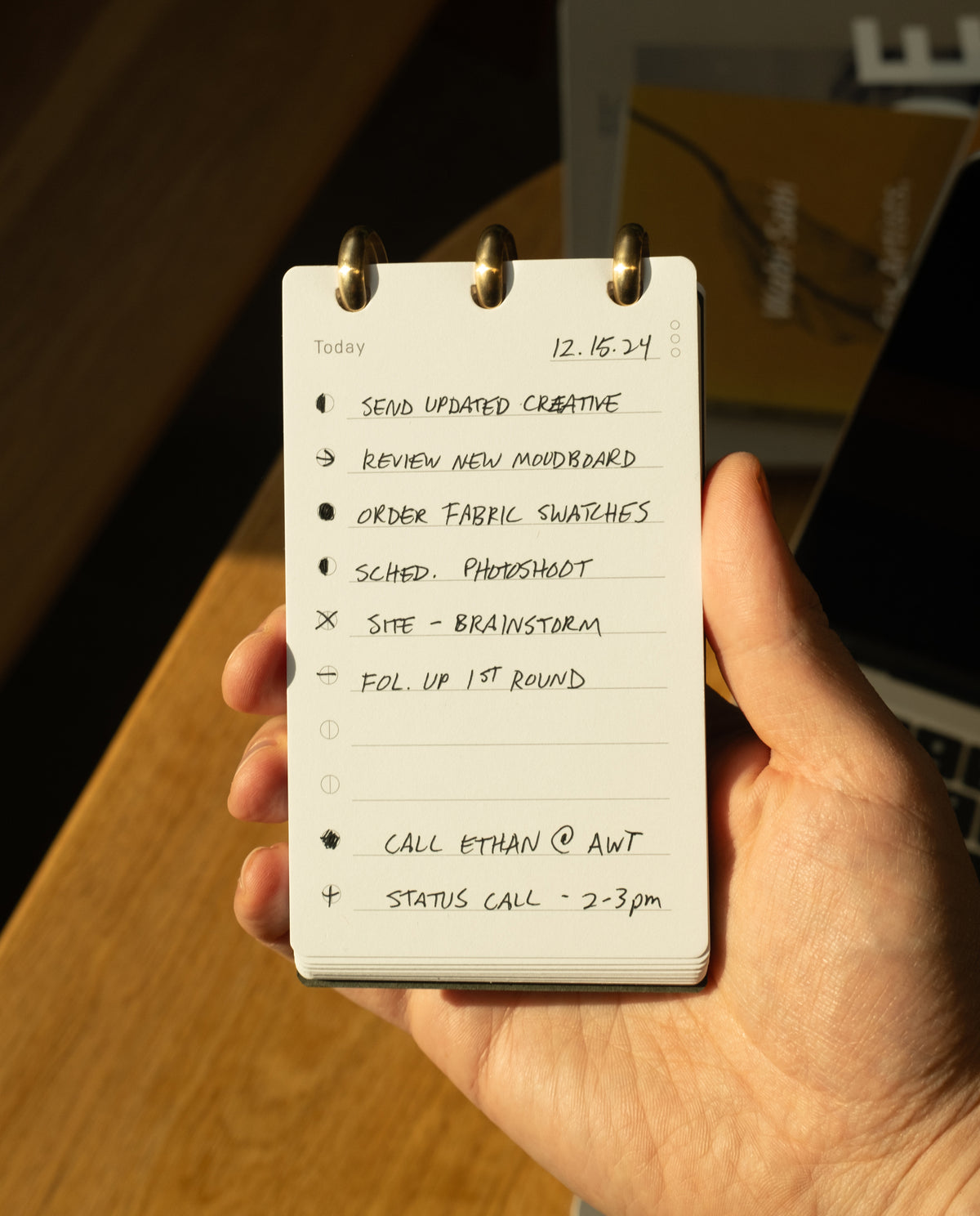 A hand holds the Discbound Analog + 3 Refills planner, open to a to-do list dated 12.16.24 with tasks like sending creative updates, reviewing a moodboard, ordering swatches, scheduling a photoshoot, brainstorming, and making calls.