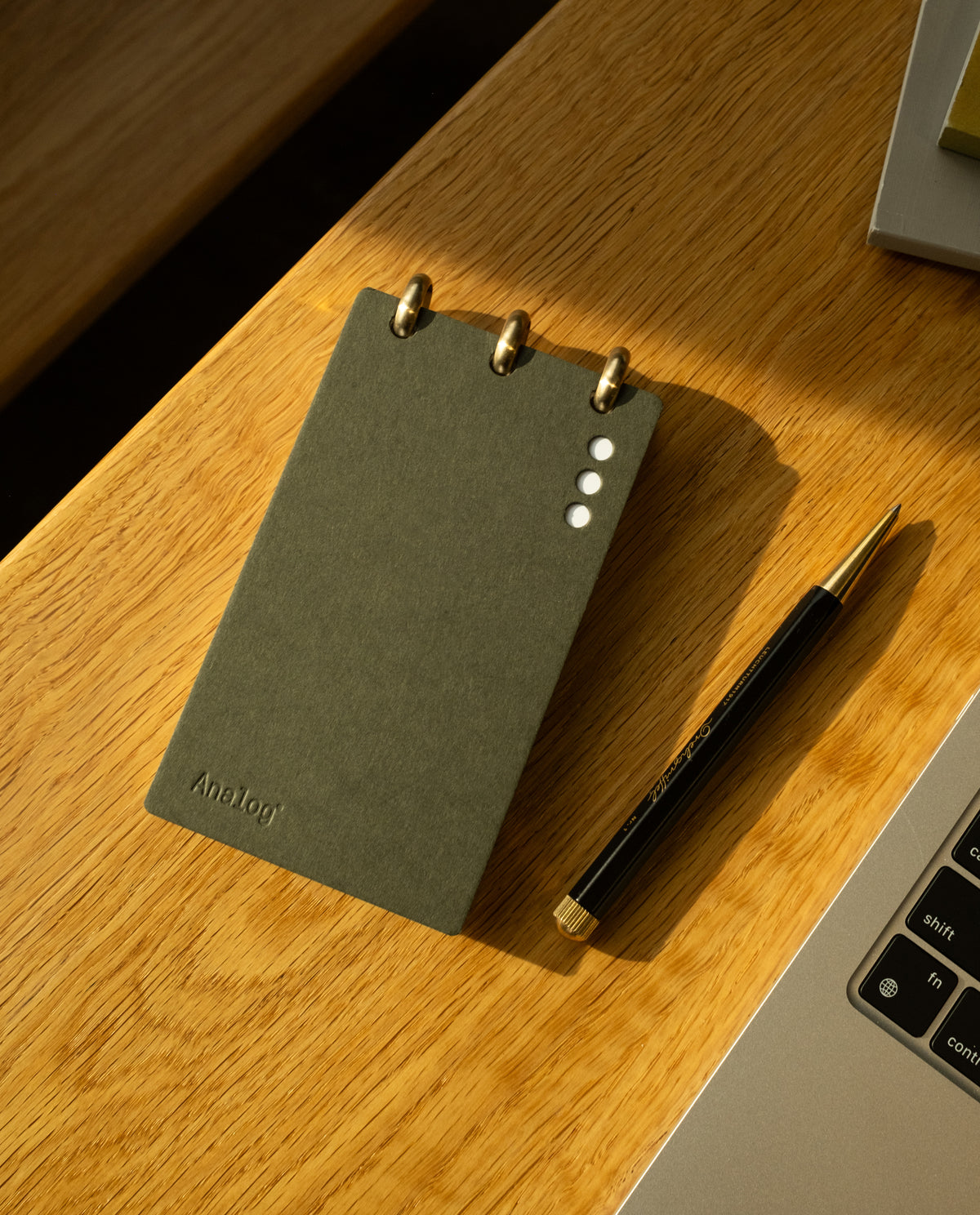 A Discbound Analog + 3 Refills notepad with brass disc binding rests on a wooden desk beside a black and gold pen and the corner of a laptop keyboard.