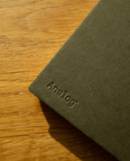 A Discbound Analog + 3 Refills notebook with brass disc binding rests on a wooden surface, soft sunlight highlighting the dark green cover and textured materials.